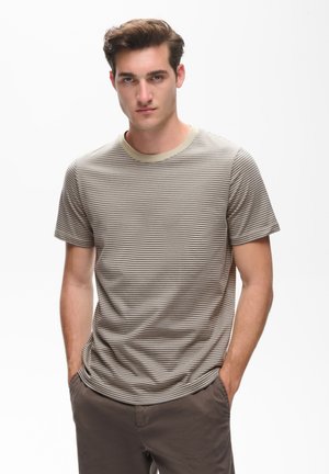 Ung mand med kort brunt hår iført en beige og brunstribet t-shirt og brune bukser, stående med hænderne i lommerne mod hvid baggrund.