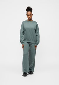 Απαλό πράσινο σετ φούτερ και παντελόνι με φαρδιά μπάτσα, κατασκευασμένα από άνετο ύφασμα. Λαιμός με στρογγυλή λαιμόκοψη, χαμηλωμένοι ώμοι και άνετη γραμμή. Μαύρα αθλητικά παπούτσια.