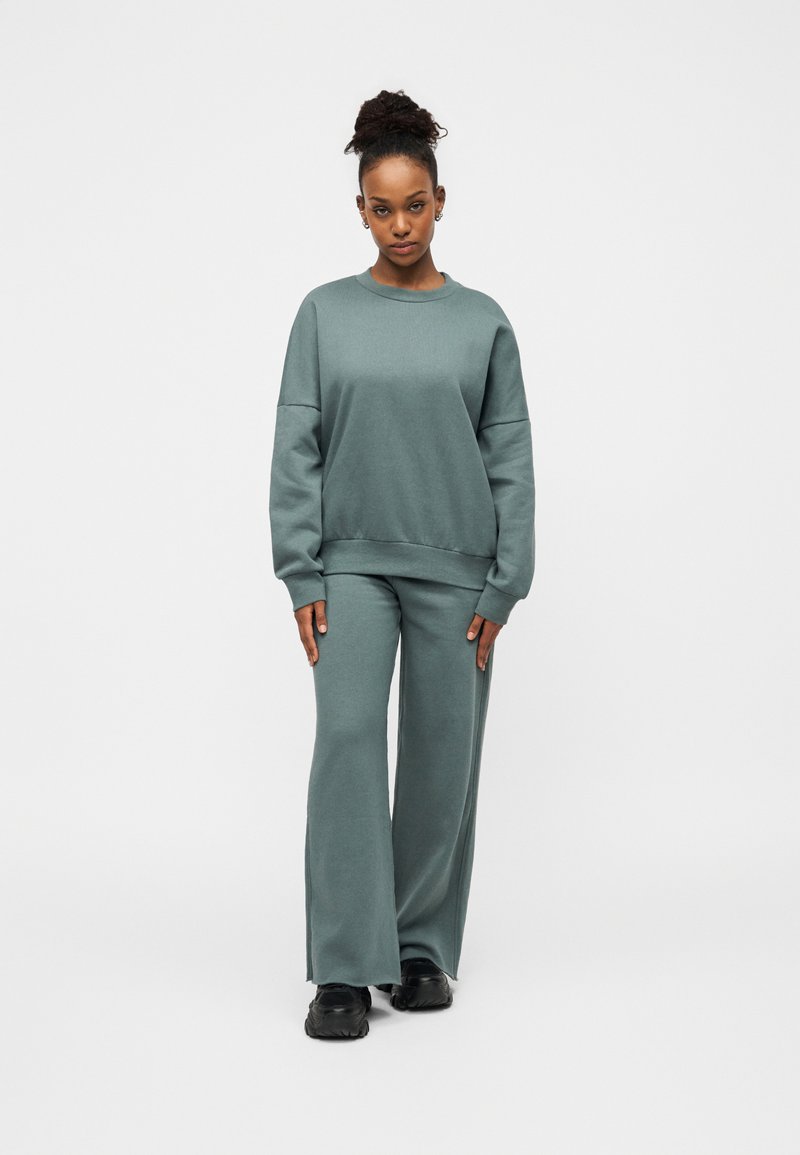 Απαλό πράσινο σετ φούτερ και παντελόνι με φαρδιά μπάτσα, κατασκευασμένα από άνετο ύφασμα. Λαιμός με στρογγυλή λαιμόκοψη, χαμηλωμένοι ώμοι και άνετη γραμμή. Μαύρα αθλητικά παπούτσια.