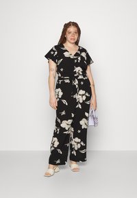 Svart blomsterjumpsuit med korte ermer, V-hals og belte i midjen. Har store hvite blomster og grønne blader, kombinert med lyse sandaler og en lavendel veske.