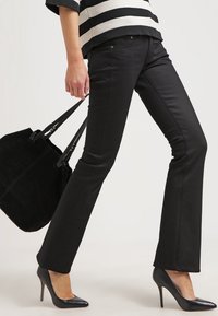 Jeans acampanados negros de denim, con una textura suave y bolsillos laterales, combinados con unos zapatos de tacón puntiagudos negros y un bolso tote negro.