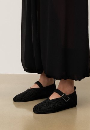 Pieds chaussés de chaussures Mary Jane noires tissées avec une boucle argentée, associés à un pantalon long noir sur un sol beige et un mur clair.
