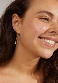 Pendientes de gota de plata con una larga cadena y un acento esférico, mostrados sobre una piel clara y un maquillaje natural.