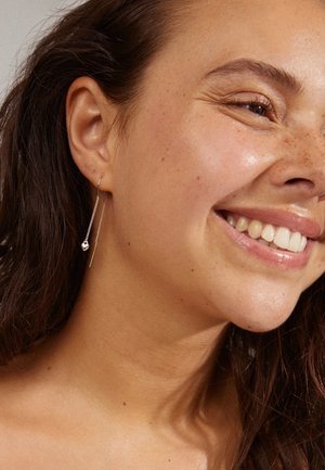 Boucles d'oreilles en argent avec une longue chaîne et un accent sphérique, mises en avant sur un teint clair et un maquillage naturel.