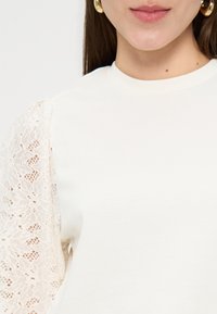 Femme portant un haut blanc avec des manches bouffantes en dentelle et des boucles d'oreilles créoles dorées, lèvres visibles, cheveux longs et bruns tombant derrière les épaules.