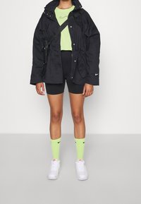 Svart oversized jacka, ljusgrön cropped top, svarta shorts, neongröna strumpor, vita sneakers. Har Nike-logotyper och justerbar rem.