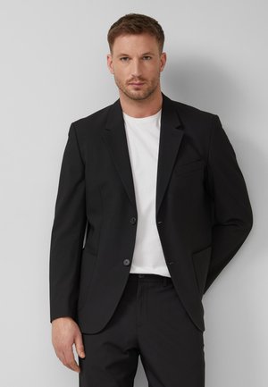 Mann trägt schwarzen Anzugjacke, schwarze Hose und weißes T-Shirt, steht vor einem einfarbigen hellgrauen Hintergrund.