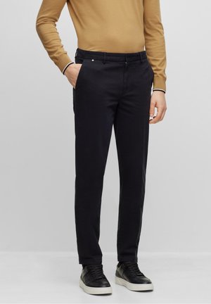Person trägt schwarze Slim-Fit-Hose, schwarze Sneakers mit weißen Sohlen und ein senffarbenes langärmeliges Oberteil, steht vor einem einfarbigen Hintergrund.