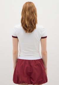 Persona con cabello rizado rojo de longitud hasta los hombros, vistiendo una camisa de manga corta blanca con ribetes burdeos y pantalones cortos burdeos, de espaldas a la cámara.