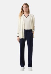Cardigan écru avec des rayures navy, superposé à un haut beige à col en V, associé à un pantalon navy et des baskets blanches, affichant une coupe décontractée.