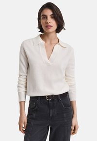 Vrouw draagt een wit, lang mouw gebreid shirt met een kraag en V-hals, gecombineerd met hoge zwarte jeans en een zwarte riem met gouden gesp.