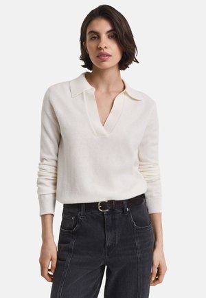 Woman wearing a white long-sleeve knit top with a collar and V-neck, paired with high-waisted black jeans and a black belt with gold buckle.