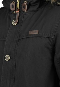 Musta takki, jossa on vetoketju- ja nappisuljenta, turkisreunuksinen huppu, nahkakoristeita ja kontrastiväriset napit teksturoidulla kankaalla.