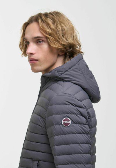 Jeune homme aux cheveux blonds mi-longs portant une veste matelassée grise avec capuche et logo Colmar sur la manche.