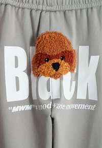 Pantalones de chándal grises con gran texto blanco que dice "Black" y "MWM x modwave movement". Un aplique de cara de animal texturizada en marrón y naranja está centrado.