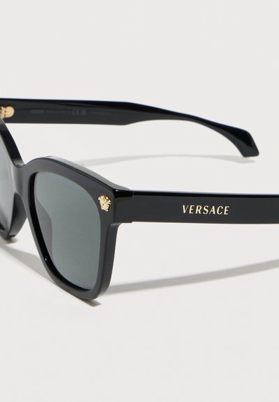 Μαύρα ορθογώνια γυαλιά ηλίου Versace με σκούρους φακούς, με μικρό χρυσό κεφάλι Μέδουσας στη γωνία και τη λέξη "VERSACE" με χρυσά γράμματα στον βραχίονα.