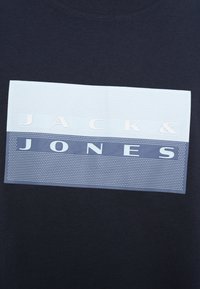 Μπλε βαμβακερό μπλουζάκι με κοντά μανίκια, διακοσμημένο με ένα ορθογώνιο σχέδιο που περιλαμβάνει το λευκό κείμενο "JACK & JONES" και μια υφασμάτινη σχεδίαση σε τρία χρωματικά τμήματα.