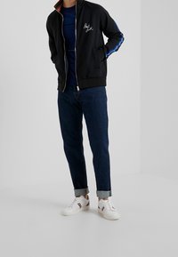 Chaqueta negra con cremallera y cuello alto, bordado de firma en blanco, rayas laterales en azul y blanco. Combinada con jeans de mezclilla oscuros enrollados y zapatillas blancas.