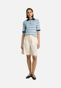 Randig blå polo t-shirt med marinblå krage, kombinerad med krämfärgade shorts. Modellen bär svarta platta skor och står mot en neutral bakgrund.
