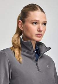 Grå zip-up tröja med ståkrage, tillverkad av mjukt tyg. Har en diskret logotyp på bröstet och en tvåfärgad design.