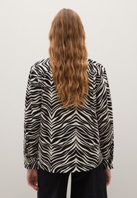 Camisa de rayas blancas y negras con un patrón de olas, de manga larga y con puños abotonados. Llevada con pantalones oscuros, vista desde la espalda.