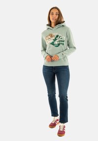Lichtgroene hoodie met grafische ontwerpen, voorzakken, geribde manchetten en zoom, gecombineerd met blauwe spijkerbroek en kastanjebruine sneakers.