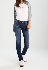 Vit t-shirt med grå ärmar som har flerfärgad "TOMMY" text, kombinerad med mörkblå jeans och vita sneakers med en beige sula.