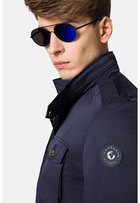 Granatowa kurtka z wysokim kołnierzem, zapięciem na guziki z przodu i kieszeniami po bokach. Posiada okrągłą naszywkę marki na lewym rękawie. Okulary przeciwsłoneczne mają niebieskie soczewki.