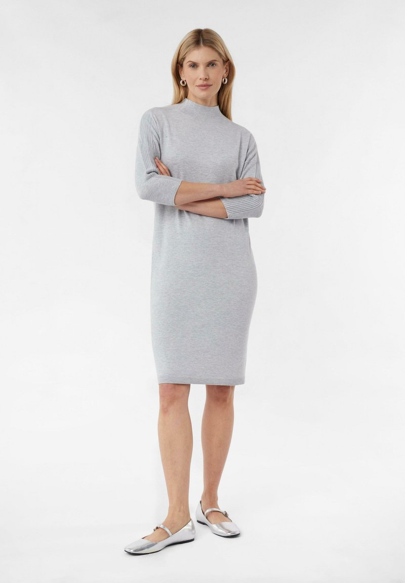 Graues gestricktes Kleid mit hohem Stehkragen, Dreiviertelärmeln und taillierter Silhouette, kombiniert mit silbernen flachen Schuhen.