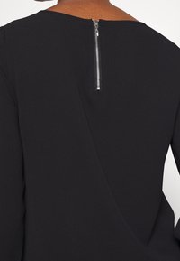 Top nero con scollatura alta e chiusura con zip sul retro. Tessuto liscio con una leggera lucentezza e un design minimalista.
