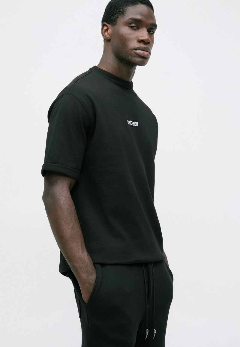 Homme avec les mains dans les poches, portant une chemise noire à manches courtes et un pantalon noir sur un fond clair uni.