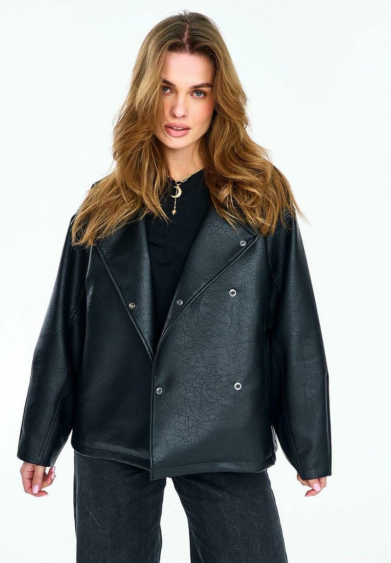 Kobieta z długimi falowanymi włosami, ubrana w oversize'ową czarną skórzaną kurtkę i czarne spodnie, zwrócona przodem na jednolitym białym tle.