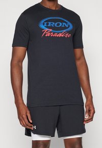 Mężczyzna ubrany w czarną koszulkę "Iron Paradise" i czarne szorty Under Armour z białymi szortami kompresyjnymi pod spodem, stojący na białym tle.