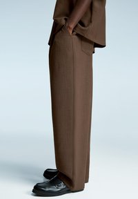 Rjave široke hlače z gladko teksturo, ki imajo stranske žepe in prilagojen kroj, v kombinaciji s sijočimi črnimi čevlji.