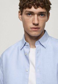 Chemise bleue clair à col, avec un léger brillant et une texture lisse ; portée sur un t-shirt blanc.