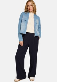 Veste en jean bleu clair avec deux poches frontales, associée à un chemisier crème texturé et un pantalon large bleu marine, portée avec des chaussures beiges sans lacets.