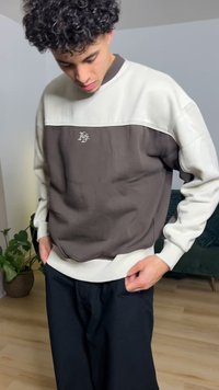 Grijs- en crèmekleurig sweatshirt met ronde hals, geribbelde manchetten en een logo op de borst, gecombineerd met gestructureerde zwarte broek. Binnenomgeving.