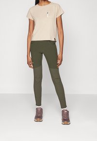 Beige t-shirt med kort ärm och ficka; gröna leggings med strukturerad fabrik och knäförstärkningar; bruna träningsskor med vita och grå detaljer.