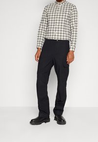 Uomo che indossa pantaloni cargo neri, stivali neri robusti e una camicia a quadri beige e grigia con bottoni, in piedi contro uno sfondo semplice.