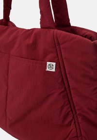 Red quilted tote bag with two sturdy handles, featuring a front pocket and a small logo patch. Material has a slightly crinkled texture.