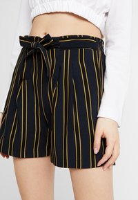 Pantalones cortos azul marino con rayas verticales amarillas, con un lazo frontal y pliegues en la cintura. Hechos de una tela suave y texturada.