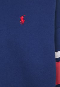 Tmavě modrý bavlněný svetr s vyšitým červeným logem. Rukávy zdobené červenobílými pruhy. Hladká textura.