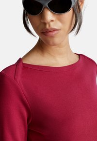 Nahaufnahme einer Person, die schwarze Sonnenbrillen und ein rotes Oberteil mit weitem Ausschnitt trägt, vor einem schlichten weißen Hintergrund.