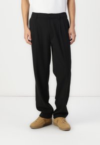 Pantaloni neri plissettati realizzati in tessuto liscio, con una vita con chiusura a bottone, abbinati a sneakers beige.
