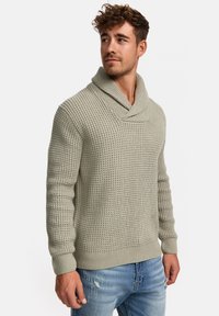 Suéter de punto en suave verde claro con un amplio cuello tipo chal, cuerpo texturizado y puños acanalados. Combinado con unos vaqueros azules desgastados.