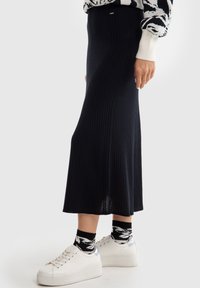 Knit black midi sijonas su brižnių tekstūra, turintis prigludusį juosmens diržą ir sklandžią siluetą. Suderintas su baltomis sportinėmis bateliais ir raštuotomis kojinėmis.