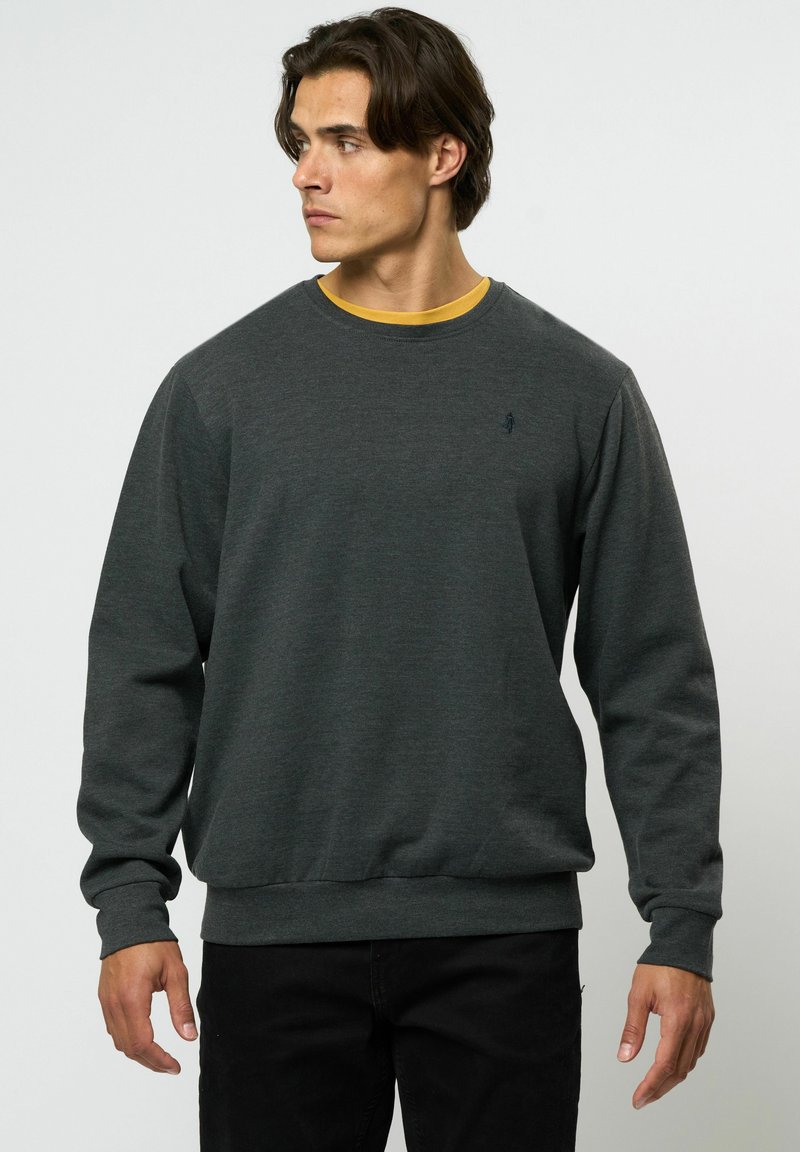 Szürke pulóver kerek nyakkivágással és hosszú ujjakkal, diszkrét logóval. Egy sárga pólóra rétegezve. Sima textúra.