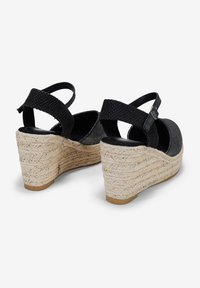 Sandales à talon noir en tissu texturé, avec des semelles style espadrille et des brides ajustables à la cheville. Elles présentent un bout arrondi et une base de couleur claire.