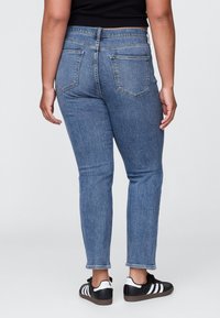 Tamsiai mėlynos spalvos tiesios kelnės iš džinso audinio, turinčios du galinius kišenes, vidutinio aukščio liemenį ir kantą šiek tiek virš kulkšnies, derinamos su juodais batais.