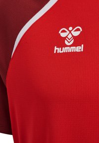 Maillot de sport rouge avec des accents bordeaux et blancs, présentant un logo de marque Hummel blanc et un emblème d'abeille sur la poitrine.